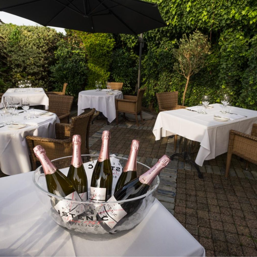 Gezellig restaurantterras met witte gedekte tafels, rieten stoelen en ISTENIČ mousserende wijn op ijs, omringd door groen.