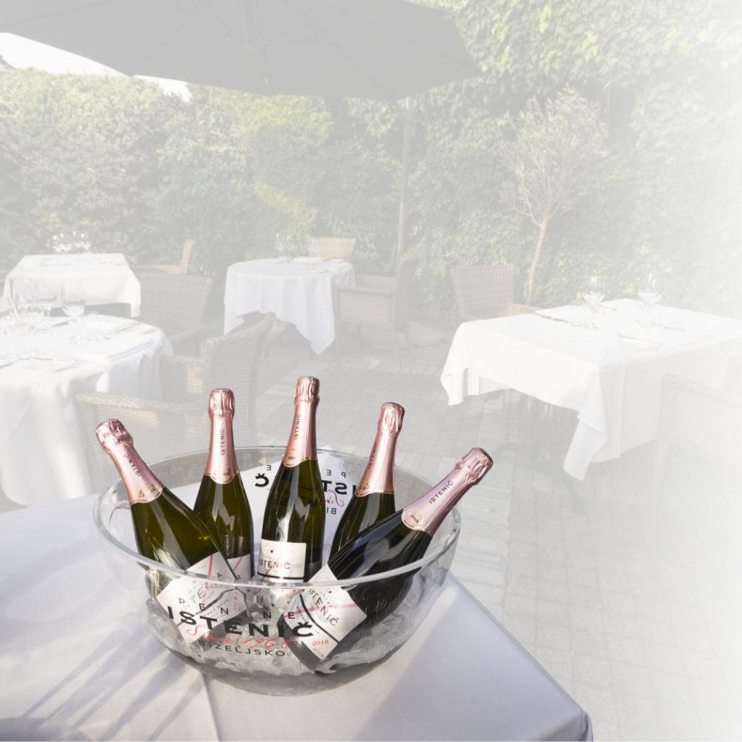 Vijf flessen Istenic mousserende wijn met roze folie in een ijsemmer op een terras.