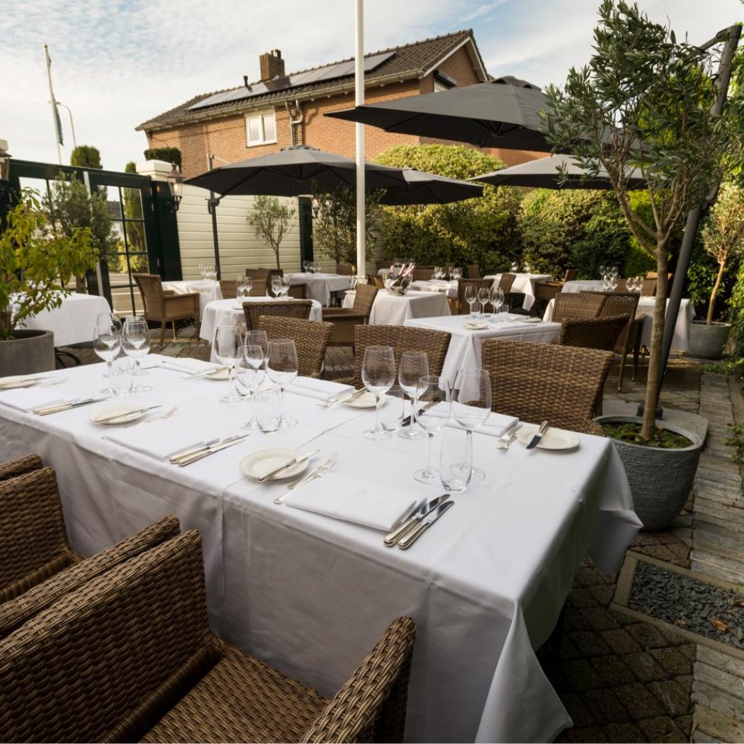 Restaurantterras met gedekte tafels, rotan stoelen, donkere parasols en groene klimop.