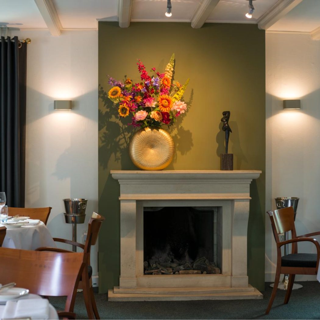 Sfeervol restaurant met een olijfgroene muur, open haard, gouden vaas met kleurrijke bloemen en gedekte tafels.
