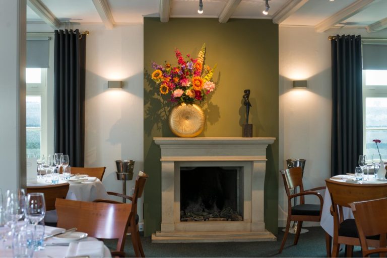 Sfeervol restaurant met een olijfgroene muur, open haard, gouden vaas met kleurrijke bloemen en gedekte tafels.