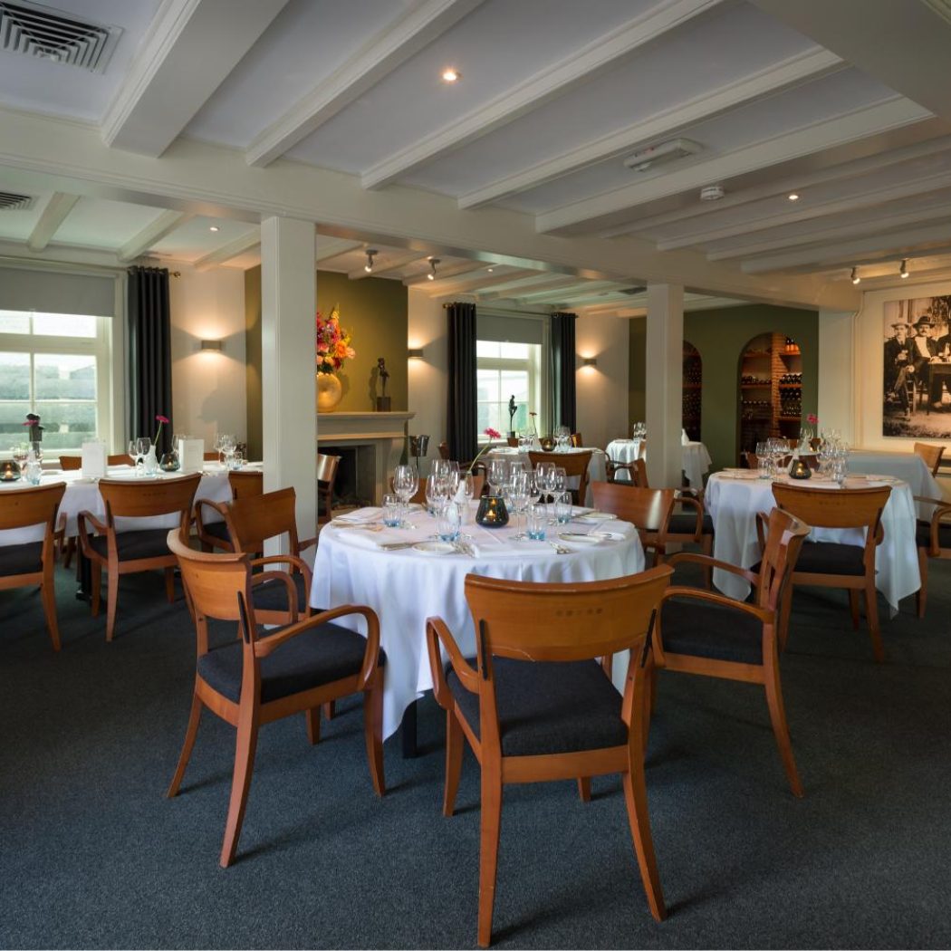 Sfeervolle restaurant dinerzaal met gedekte tafels, houten stoelen, ramen en een grote zwart-wit groepsfoto aan de muur.