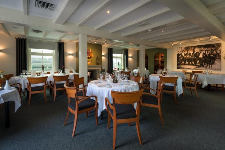 Sfeervolle restaurant dinerzaal met gedekte tafels, houten stoelen, ramen en een grote zwart-wit groepsfoto aan de muur.
