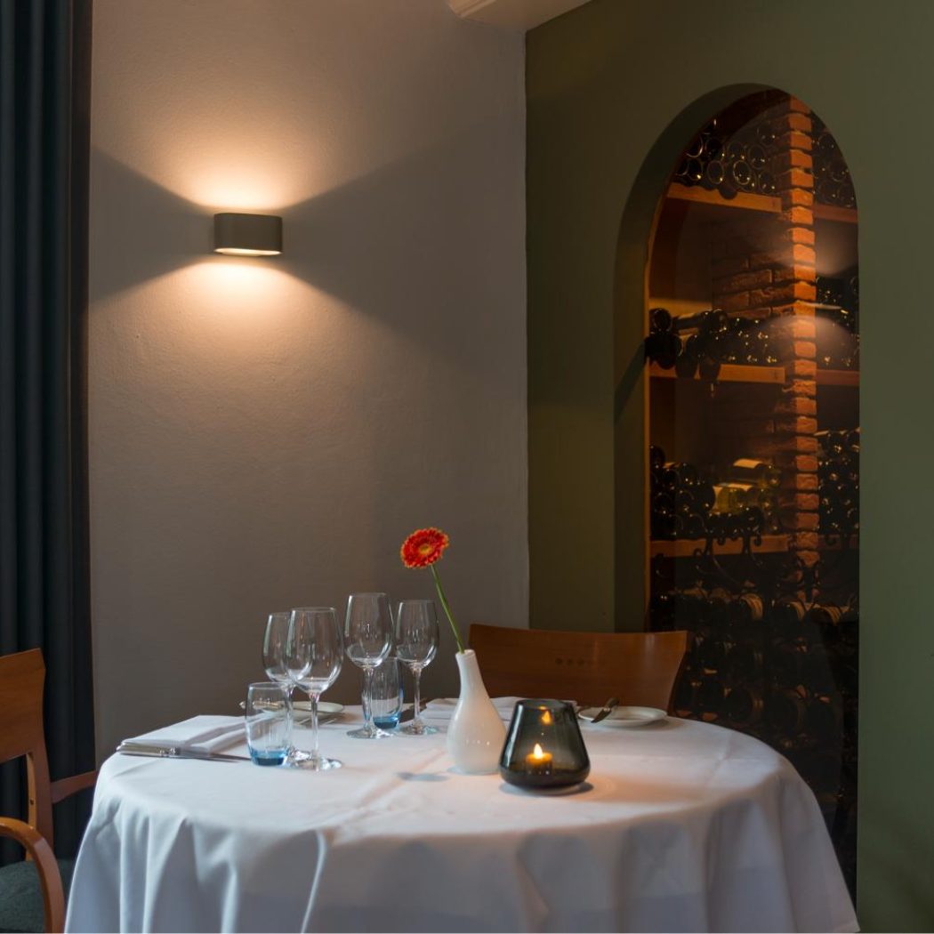 Restauranttafel met wit tafelkleed, rode bloem en kaarslicht, grenzend aan wijnnissen en een raam.