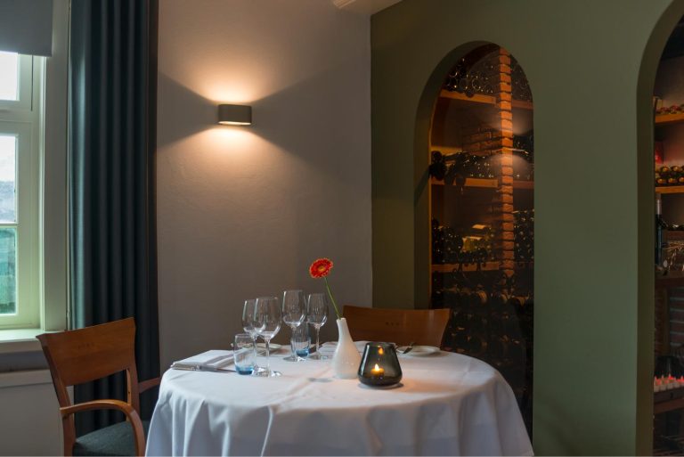 Restauranttafel met wit tafelkleed, rode bloem en kaarslicht, grenzend aan wijnnissen en een raam.