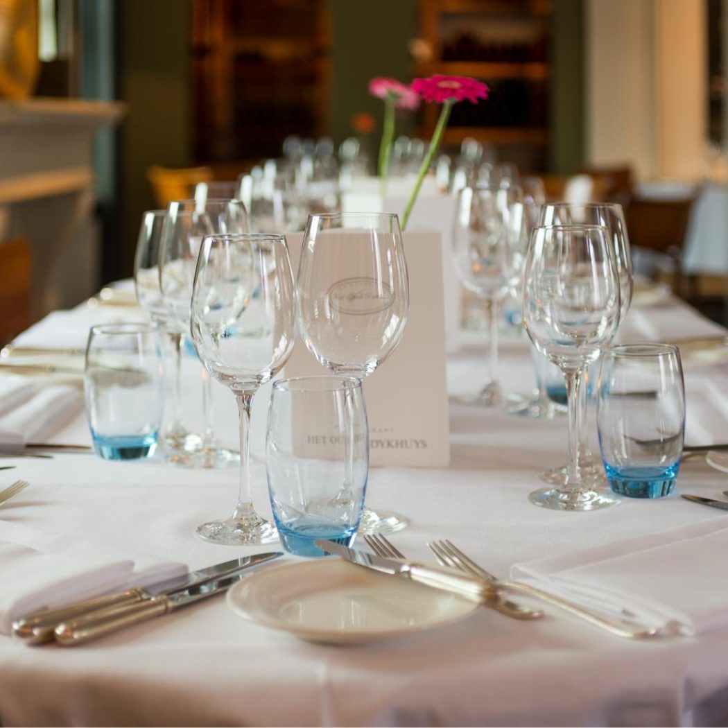 Lange restauranttafel gedekt met witte tafelkleden, glazen met blauwe voet, zilver bestek, een roze bloem en een kaartje: HET OU DYKHUYS.