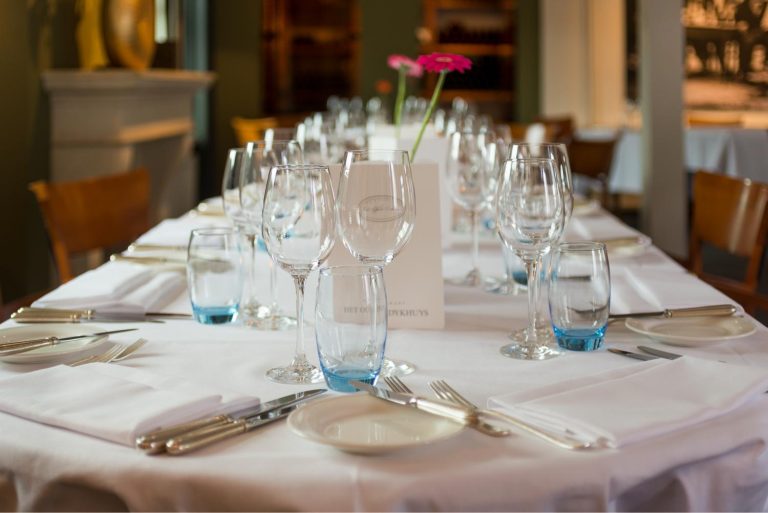 Lange restauranttafel gedekt met witte tafelkleden, glazen met blauwe voet, zilver bestek, een roze bloem en een kaartje: HET OU DYKHUYS.