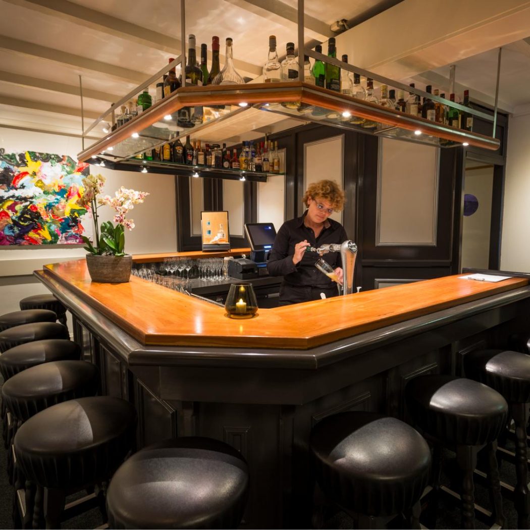 Een barman met krullend haar tapt een biertje aan een houten bar met leren krukken, een orchidee en abstracte kunst.