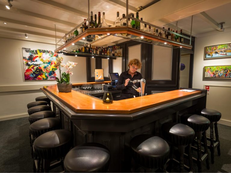 Een barman met krullend haar tapt een biertje aan een houten bar met leren krukken, een orchidee en abstracte kunst.