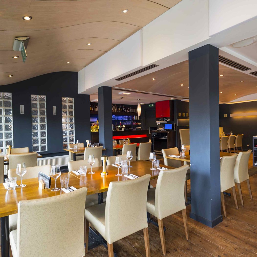 Modern restaurantinterieur met opgedekte houten tafels, beige stoelen, donkerblauwe muren, een golvend plafond en een bar.