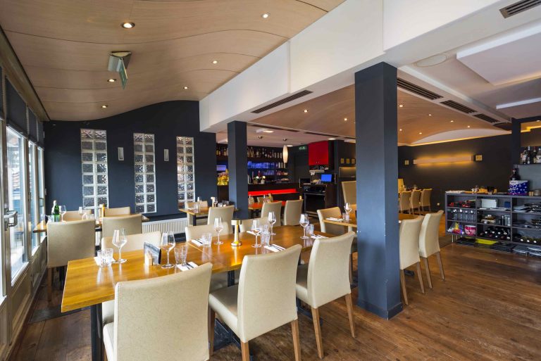 Modern restaurantinterieur met opgedekte houten tafels, beige stoelen, donkerblauwe muren, een golvend plafond en een bar.