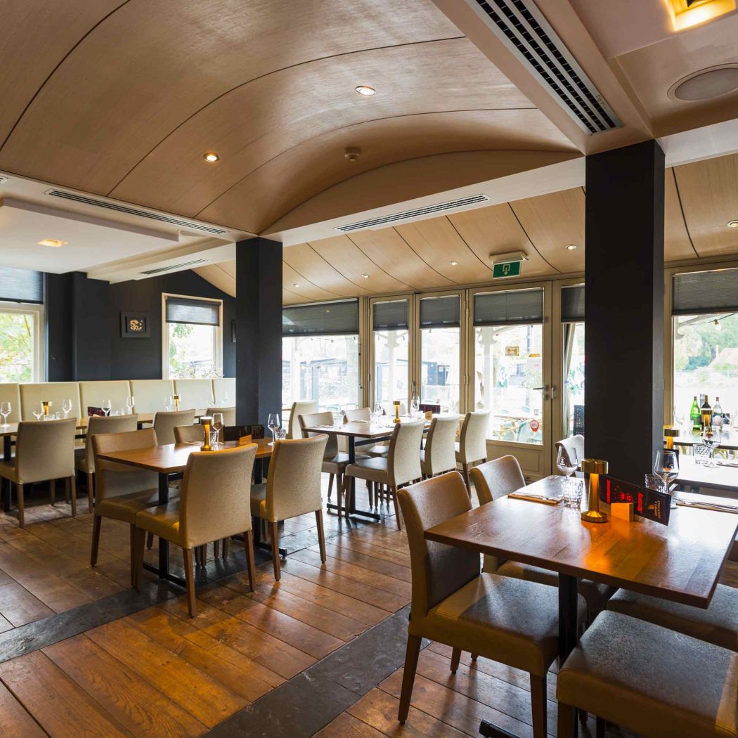 Sfeervol restaurant met houten tafels, stoelen, een gebogen houten plafond en grote ramen.