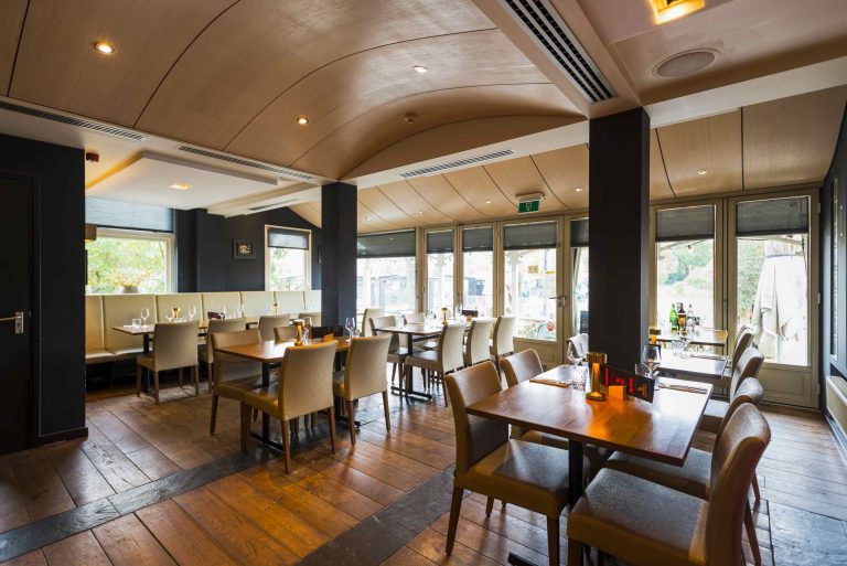 Sfeervol restaurant met houten tafels, stoelen, een gebogen houten plafond en grote ramen.