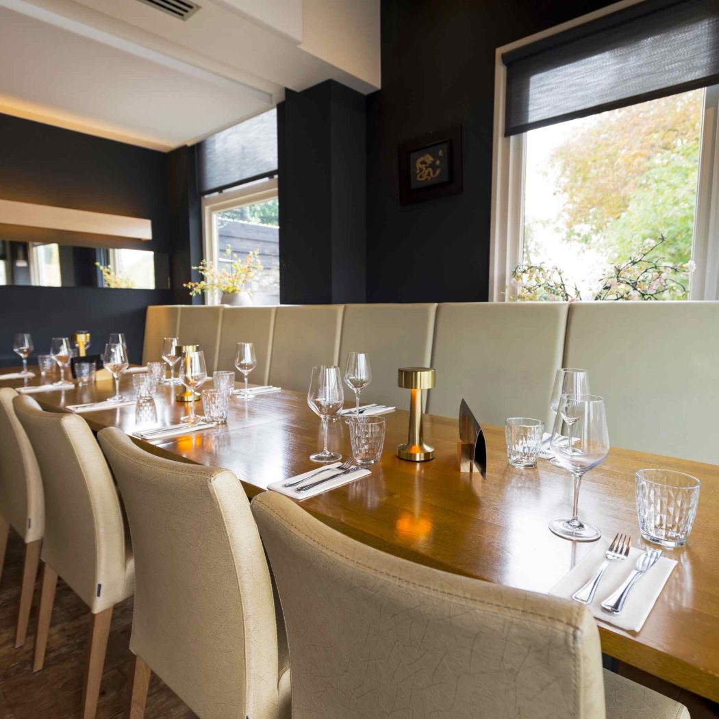 Lange houten restauranttafel met beige stoelen en bank, gedekt met servies en gouden tafellampen, bij een raam.
