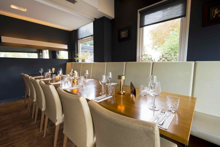 Lange houten restauranttafel met beige stoelen en bank, gedekt met servies en gouden tafellampen, bij een raam.