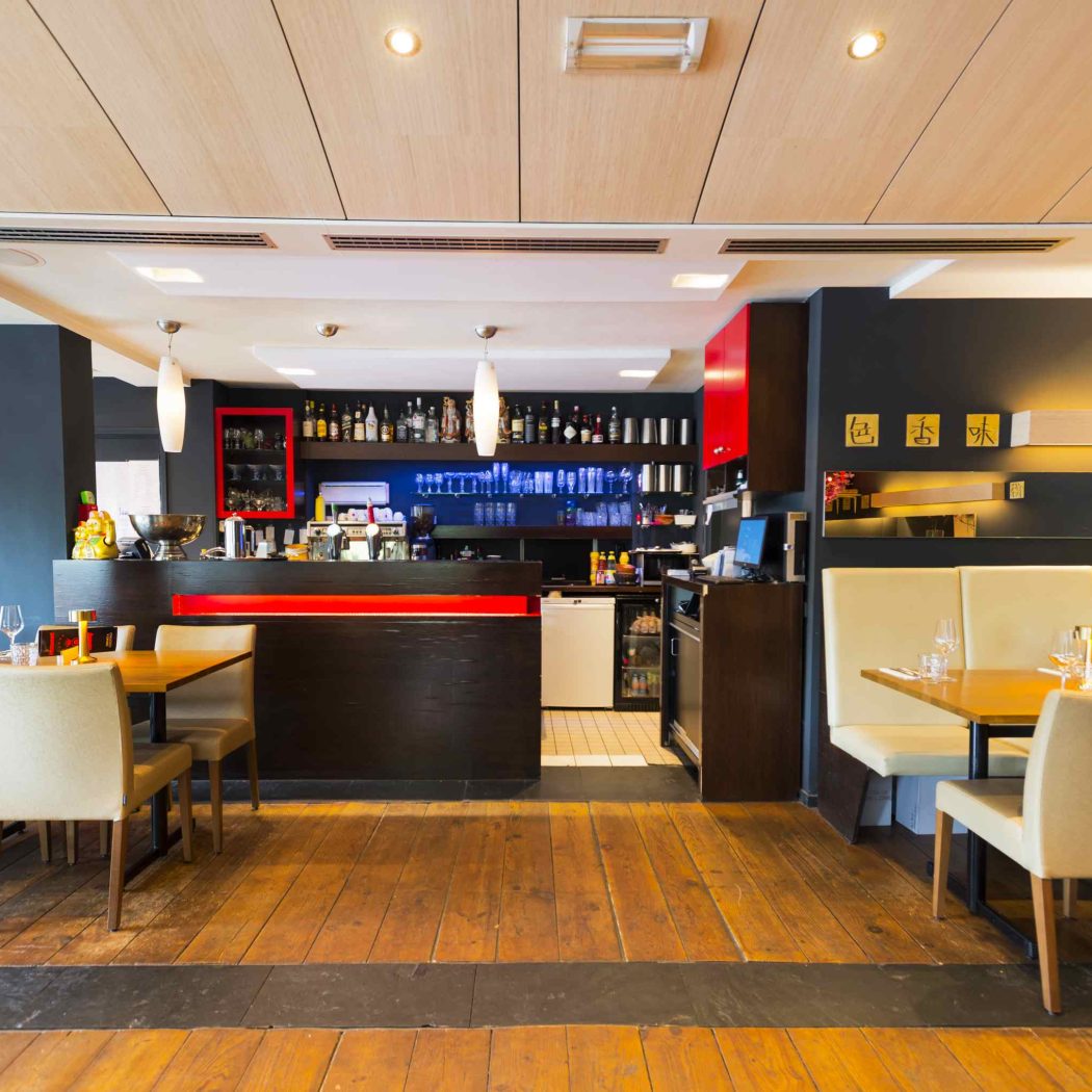 Modern restaurantinterieur met een donkere bar met rode accenten, houten vloer, beige stoelen en een flessenwand.