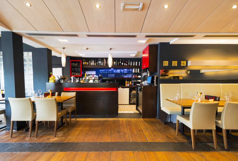 Modern restaurantinterieur met een donkere bar met rode accenten, houten vloer, beige stoelen en een flessenwand.