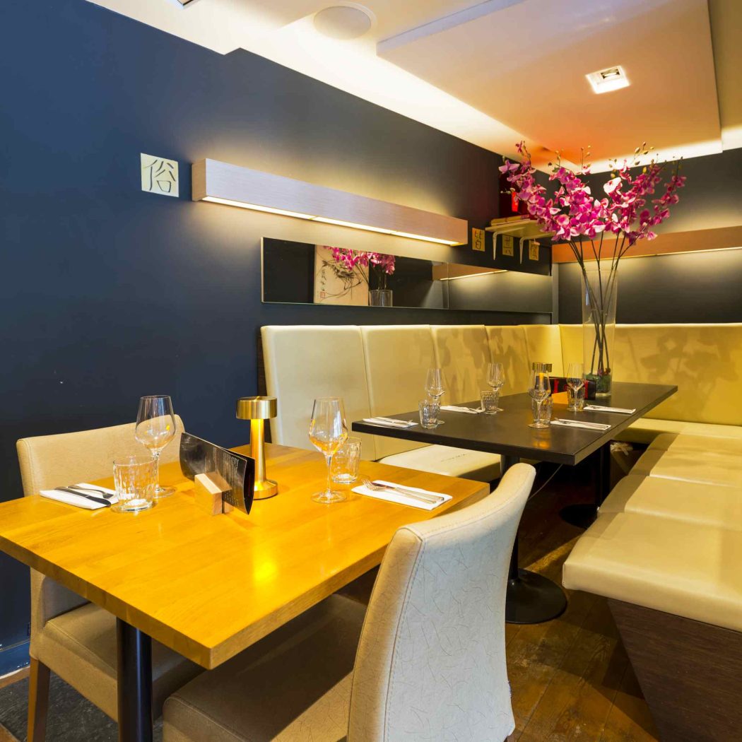 Restaurantinterieur met donkerblauwe wanden, houten tafels, crèmekleurige banken en een vaas met roze bloemen.