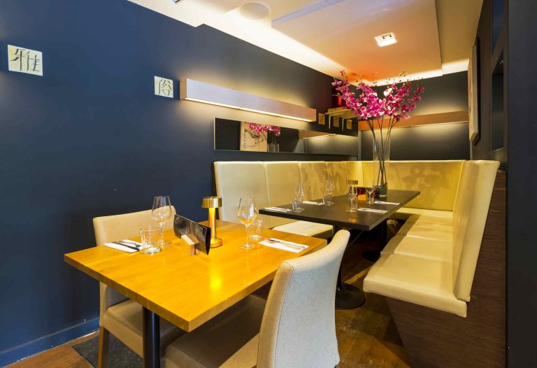 Restaurantinterieur met donkerblauwe wanden, houten tafels, crèmekleurige banken en een vaas met roze bloemen.