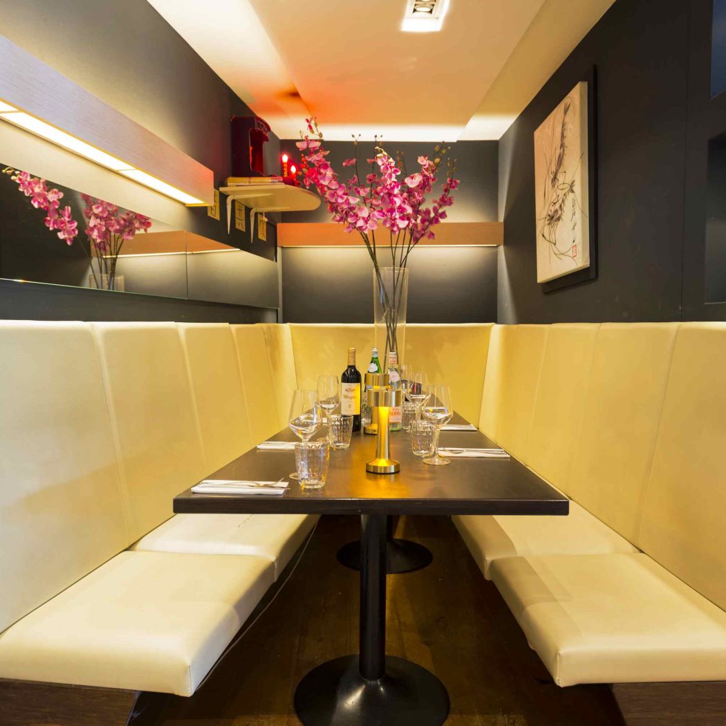 Een crèmekleurige banketbank en donkerhouten tafel in een modern restaurant, gedekt met glazen en roze bloemen.