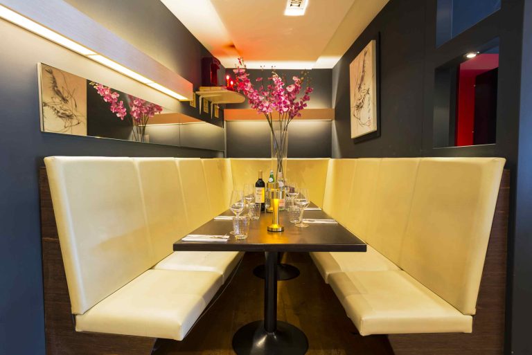Een crèmekleurige banketbank en donkerhouten tafel in een modern restaurant, gedekt met glazen en roze bloemen.