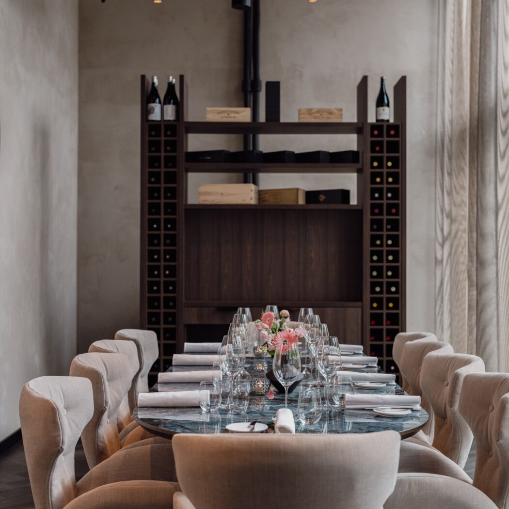 Gedekte lange marmeren tafel met roze bloemen, beige eetkamerstoelen en een wijnrek in een restaurant.