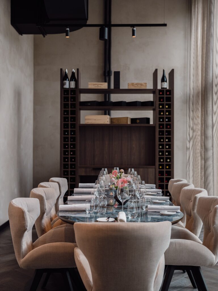 Gedekte lange marmeren tafel met roze bloemen, beige eetkamerstoelen en een wijnrek in een restaurant.