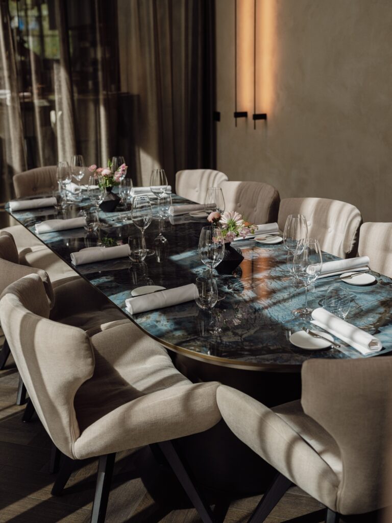Lange, elegant gedekte dinertafel met blauw marmeren blad, beige stoelen en roze bloemstukken.