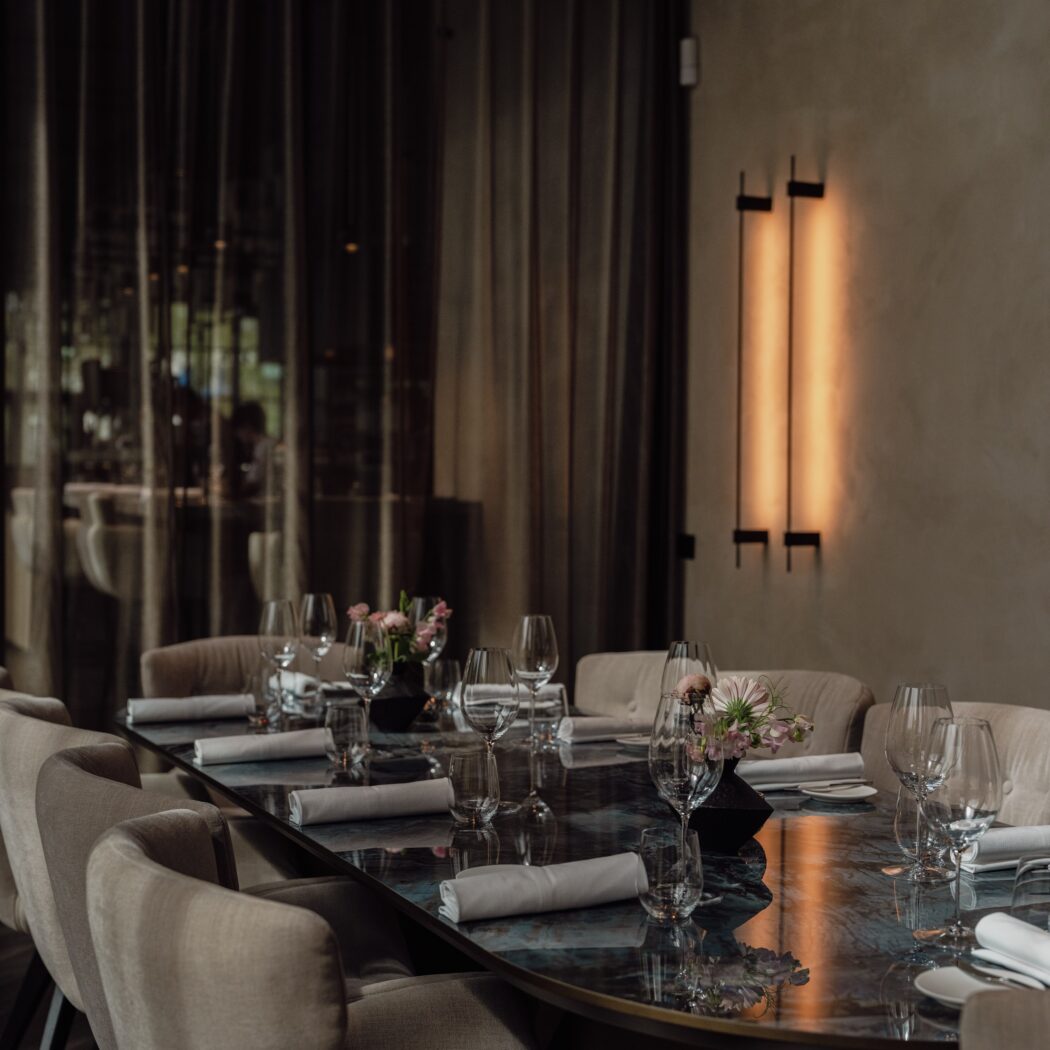 Lange, donkere, glanzende ovale tafel elegant gedekt met beige stoelen en bloemstukjes in een sfeervol restaurant.