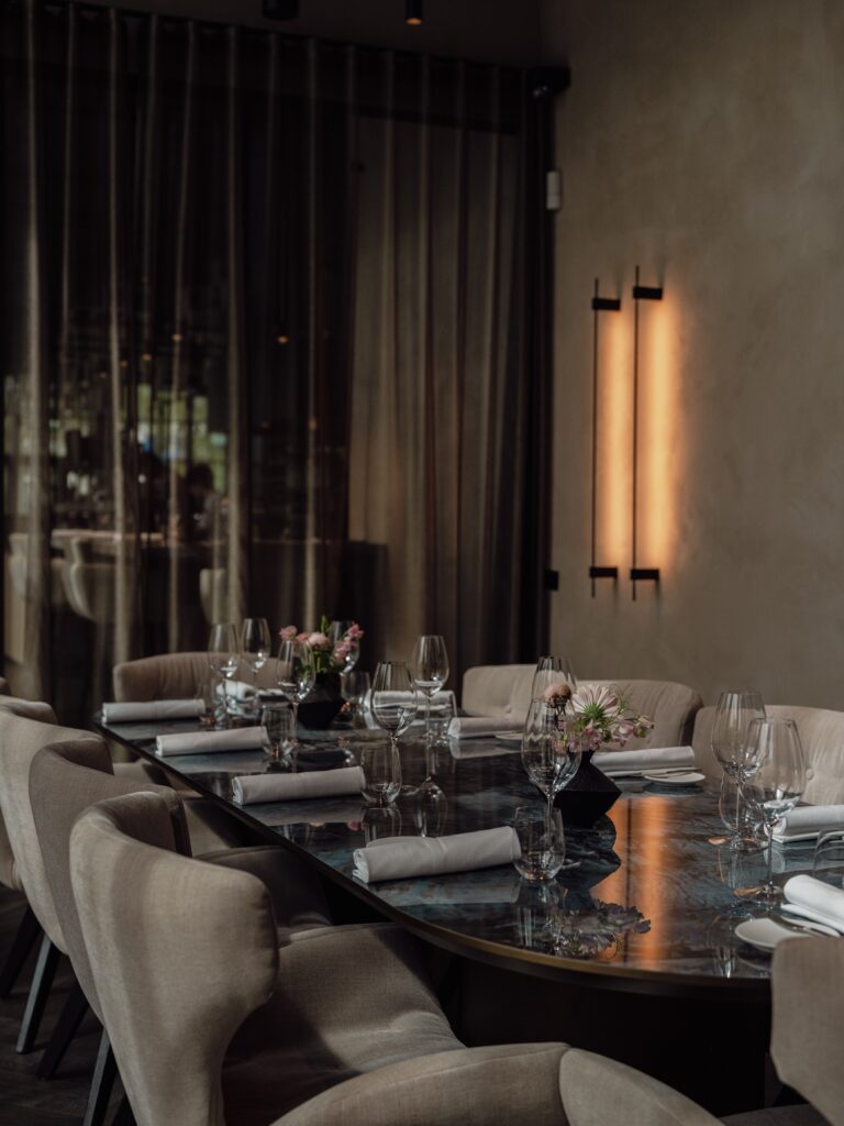 Lange, donkere, glanzende ovale tafel elegant gedekt met beige stoelen en bloemstukjes in een sfeervol restaurant.