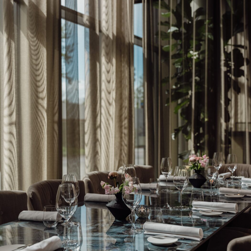 Lange, donkere marmeren eettafel met glazen, servetten en bloemen in een elegante eetzaal met grote ramen.