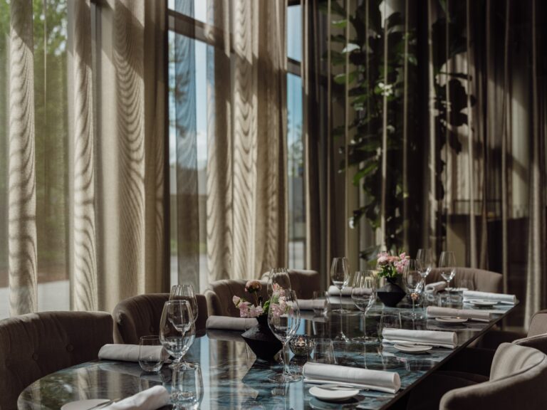 Lange, donkere marmeren eettafel met glazen, servetten en bloemen in een elegante eetzaal met grote ramen.