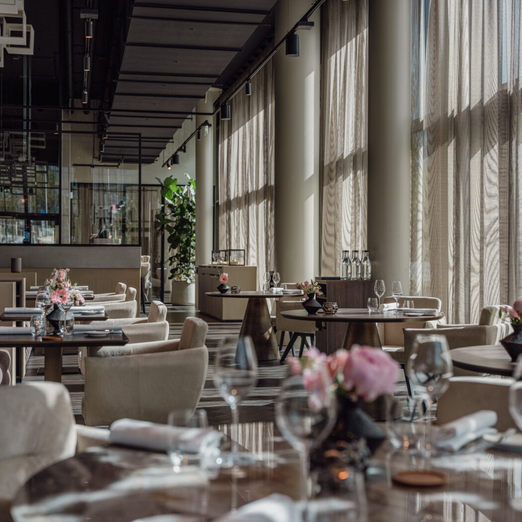 Elegant, modern restaurant interieur met gedekte tafels, roze bloemen en zonlicht door hoge ramen.