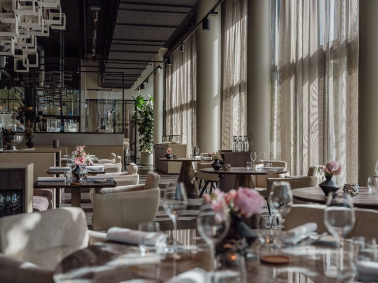 Elegant, modern restaurant interieur met gedekte tafels, roze bloemen en zonlicht door hoge ramen.