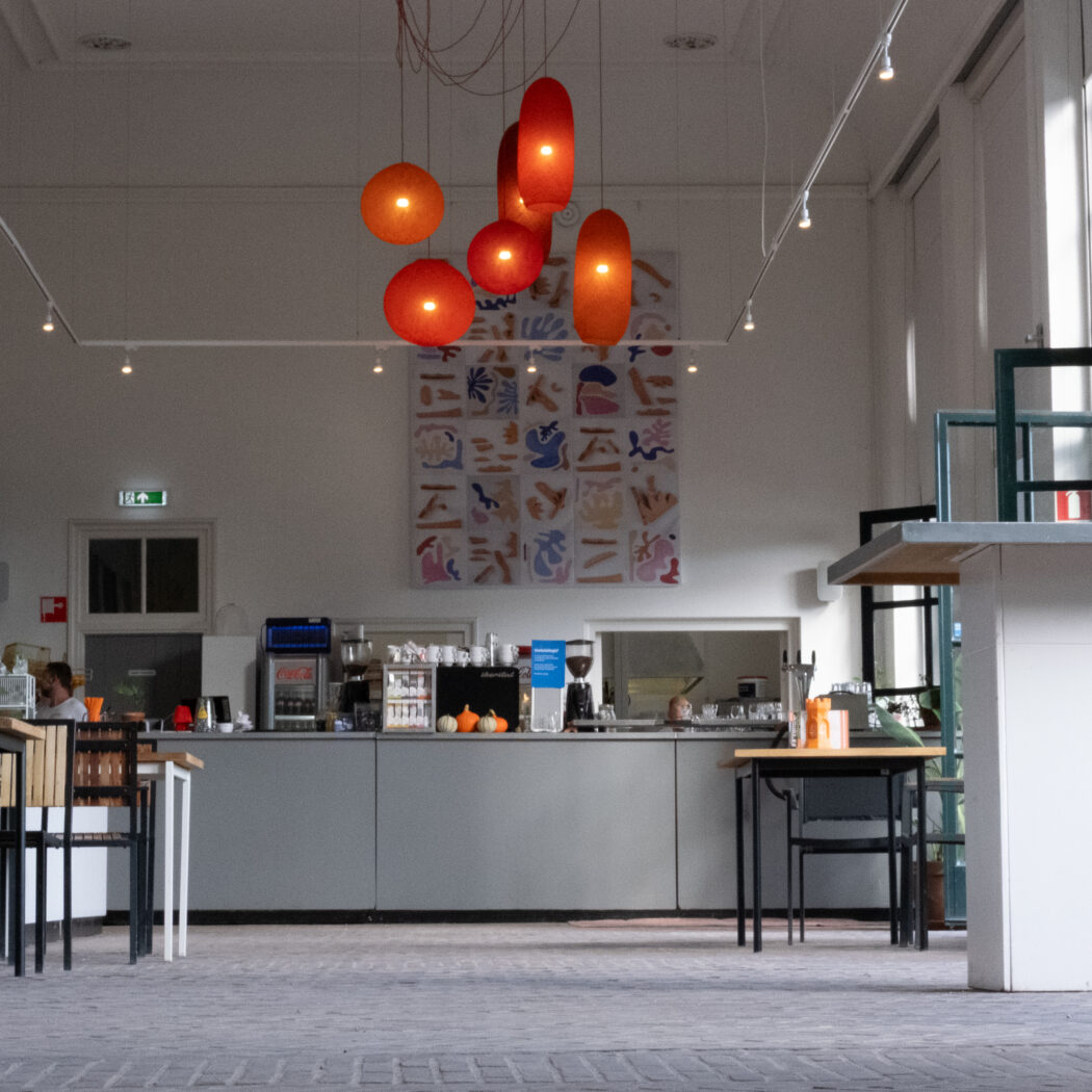 Sfeervol modern café met oranje hanglampen boven een lange grijze balie, abstracte kunst en grote glazen deuren.