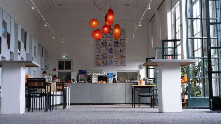 Sfeervol modern café met oranje hanglampen boven een lange grijze balie, abstracte kunst en grote glazen deuren.