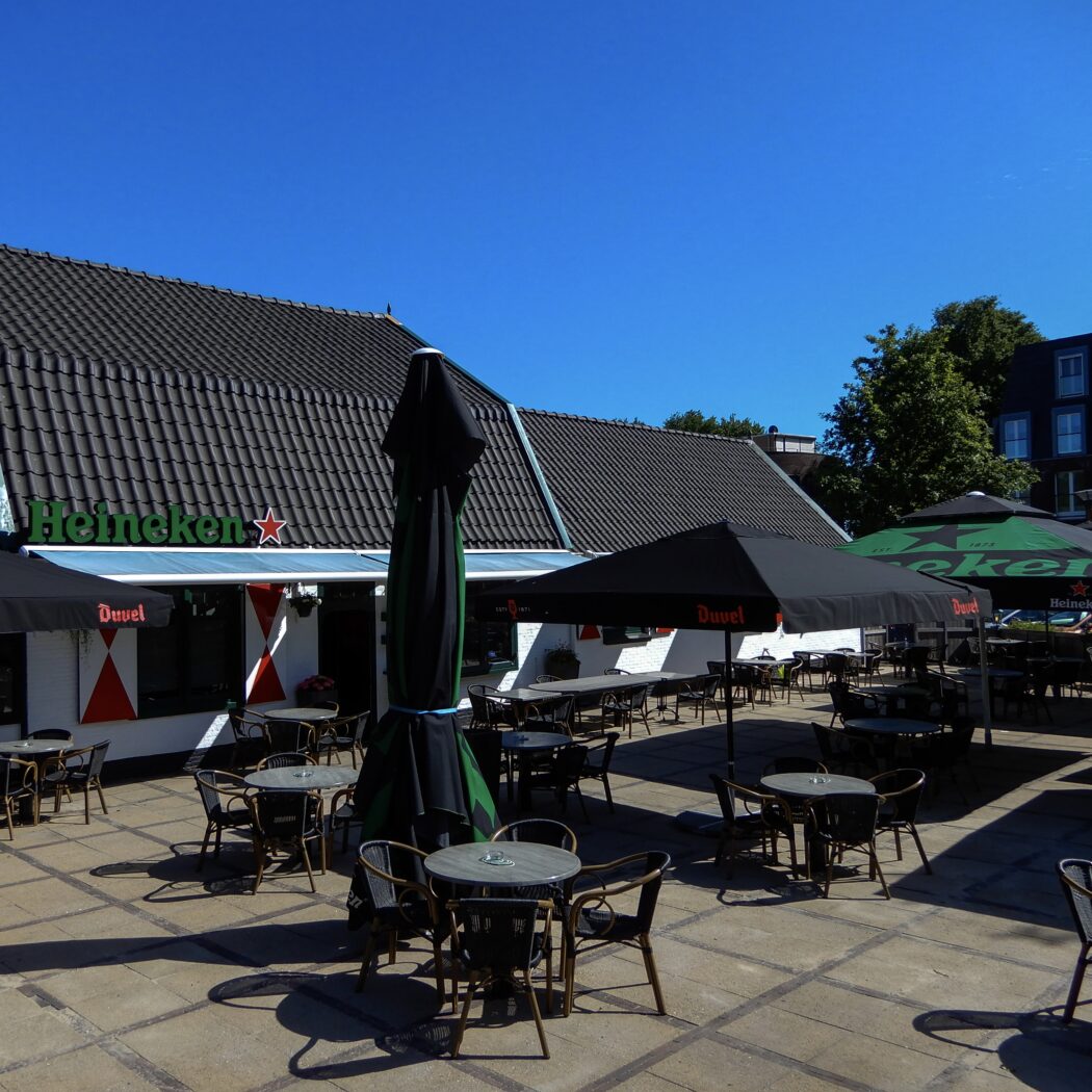Caféterras met Heineken-logo op gevel, zwarte en groene parasols van Duvel en Heineken, lege tafels.
