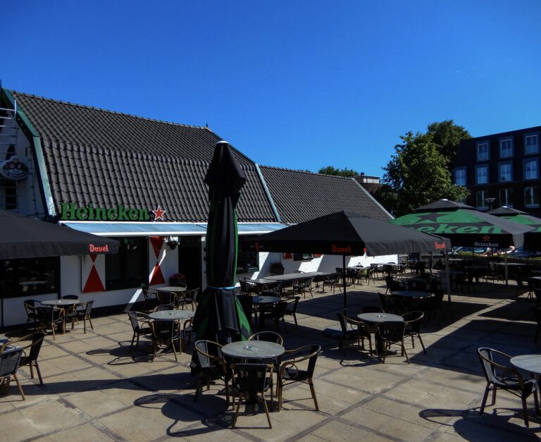 Caféterras met Heineken-logo op gevel, zwarte en groene parasols van Duvel en Heineken, lege tafels.