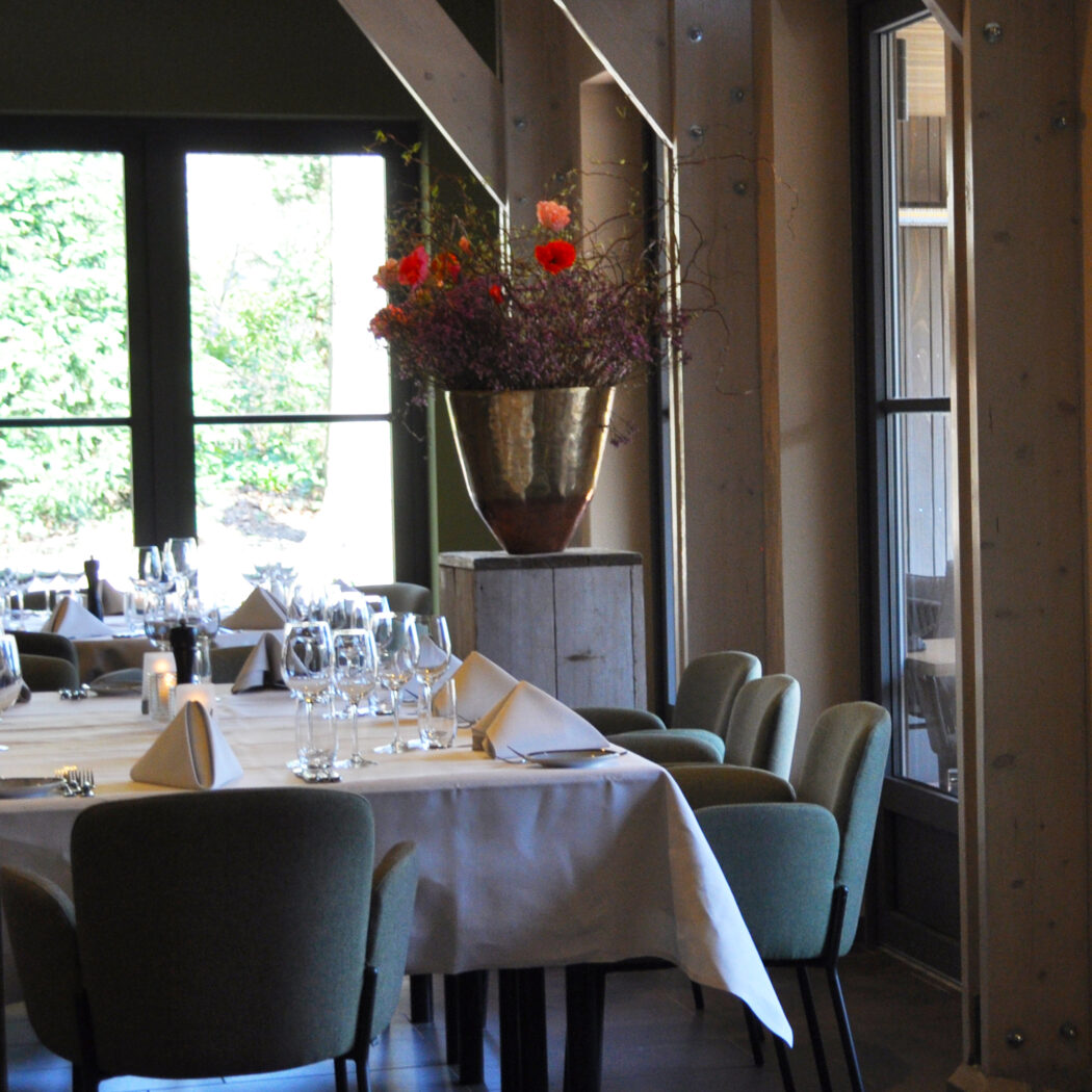 Gedekte tafel in een restaurant met groene stoelen, wijnglazen en een groot bloemstuk in een gouden vaas bij de ramen.