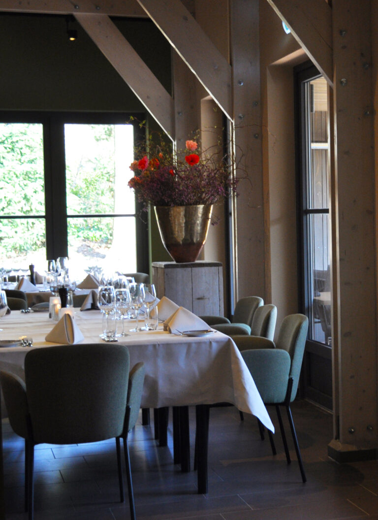 Gedekte tafel in een restaurant met groene stoelen, wijnglazen en een groot bloemstuk in een gouden vaas bij de ramen.