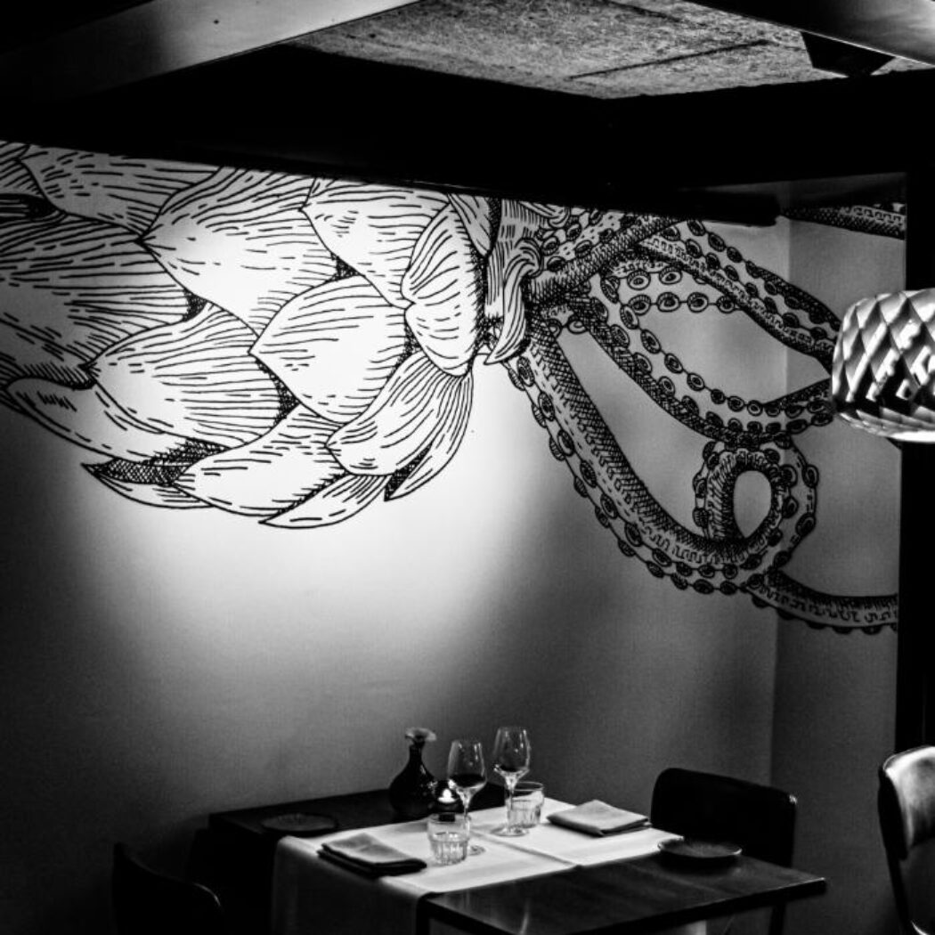 Sfeervolle gedekte tafel in een restaurant, met een muurschildering van een artisjok en octopus.