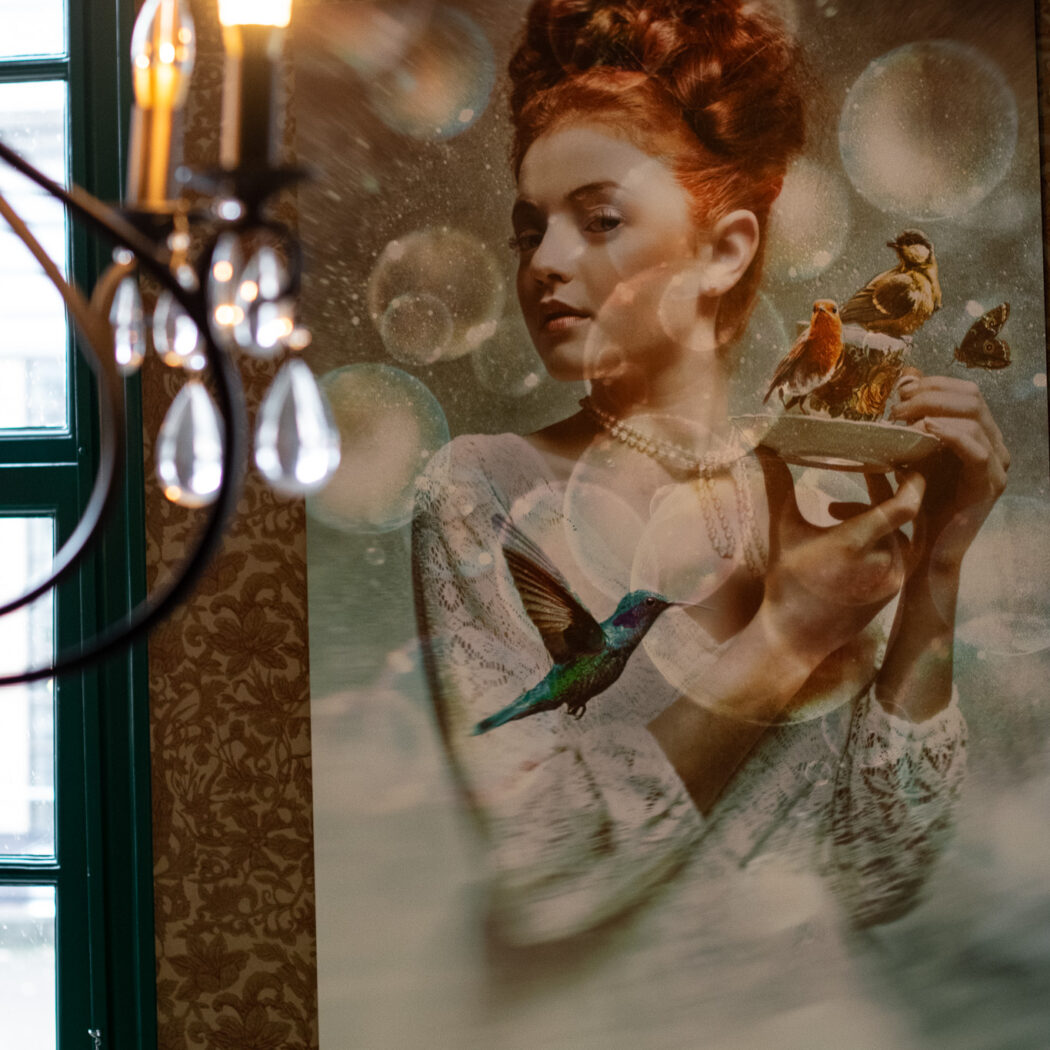 Groot kunstwerk aan de muur: een roodharige vrouw met parelketting toont vogels op een schotel, met een kolibrie.