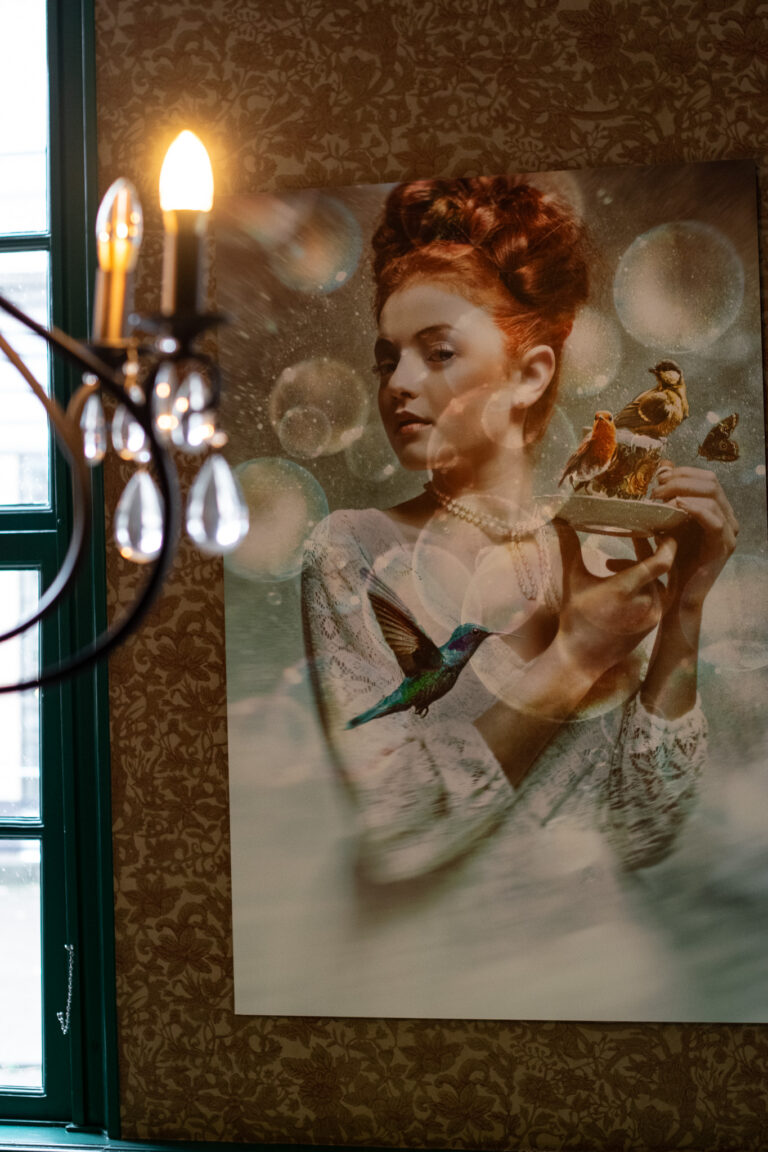 Groot kunstwerk aan de muur: een roodharige vrouw met parelketting toont vogels op een schotel, met een kolibrie.