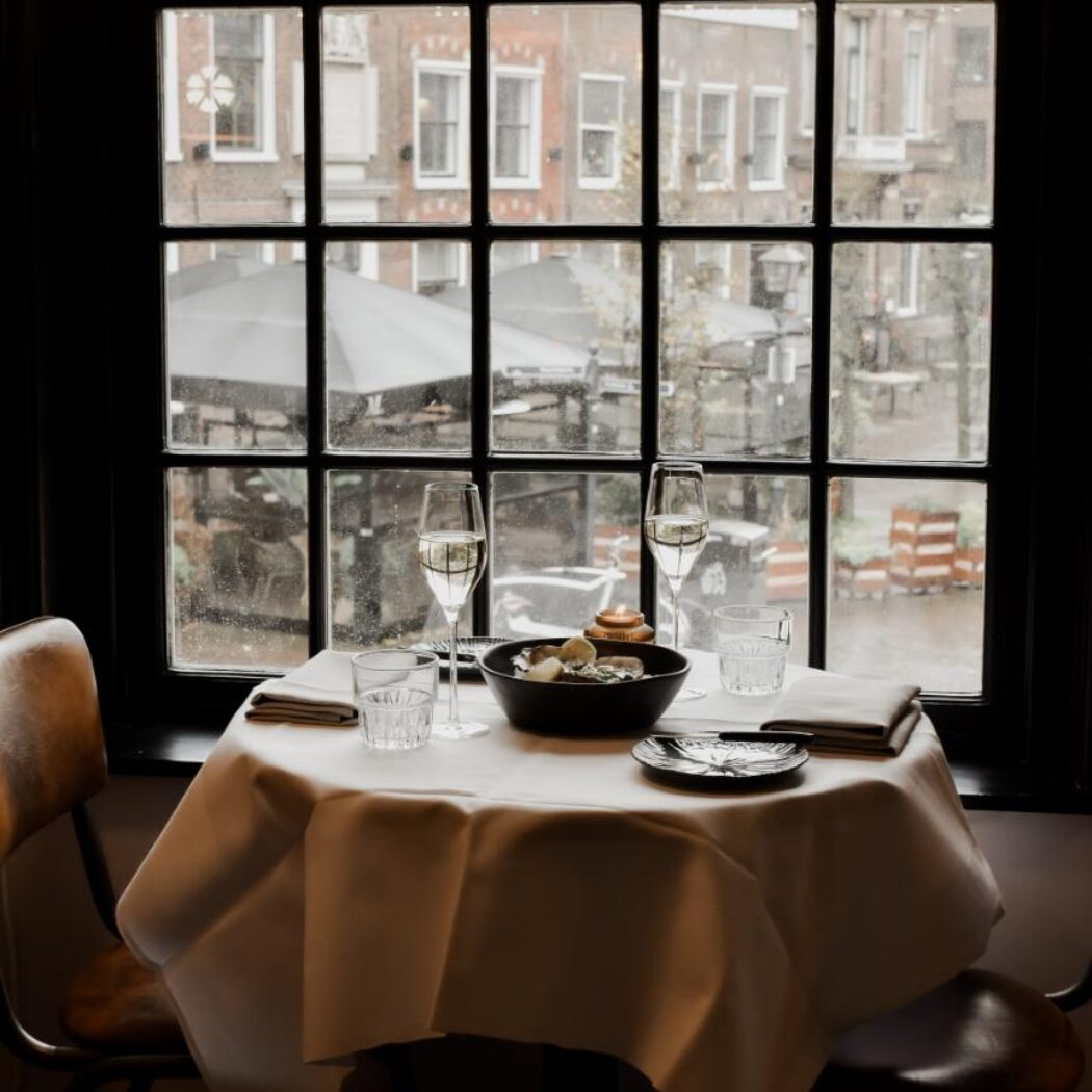 Gedekte tafel voor twee met wijnglazen, borden en oesters, bij een raam met uitzicht op stadsgebouwen.