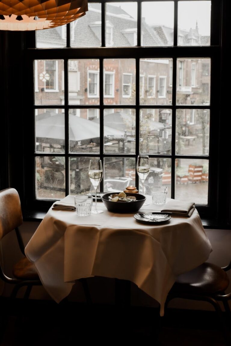 Gedekte tafel voor twee met wijnglazen, borden en oesters, bij een raam met uitzicht op stadsgebouwen.