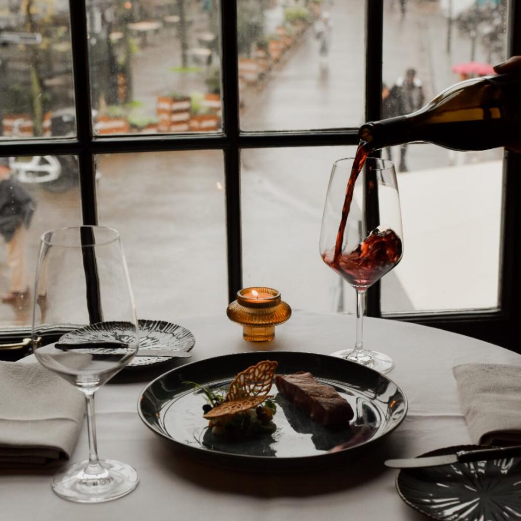 Rode wijn wordt geschonken in een glas op een restauranttafel met een biefstuk, kaars en raamuitzicht.