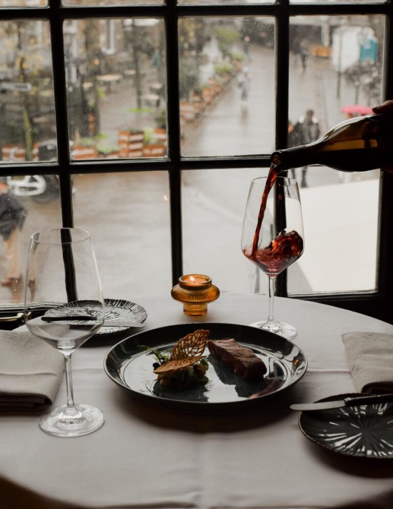 Rode wijn wordt geschonken in een glas op een restauranttafel met een biefstuk, kaars en raamuitzicht.
