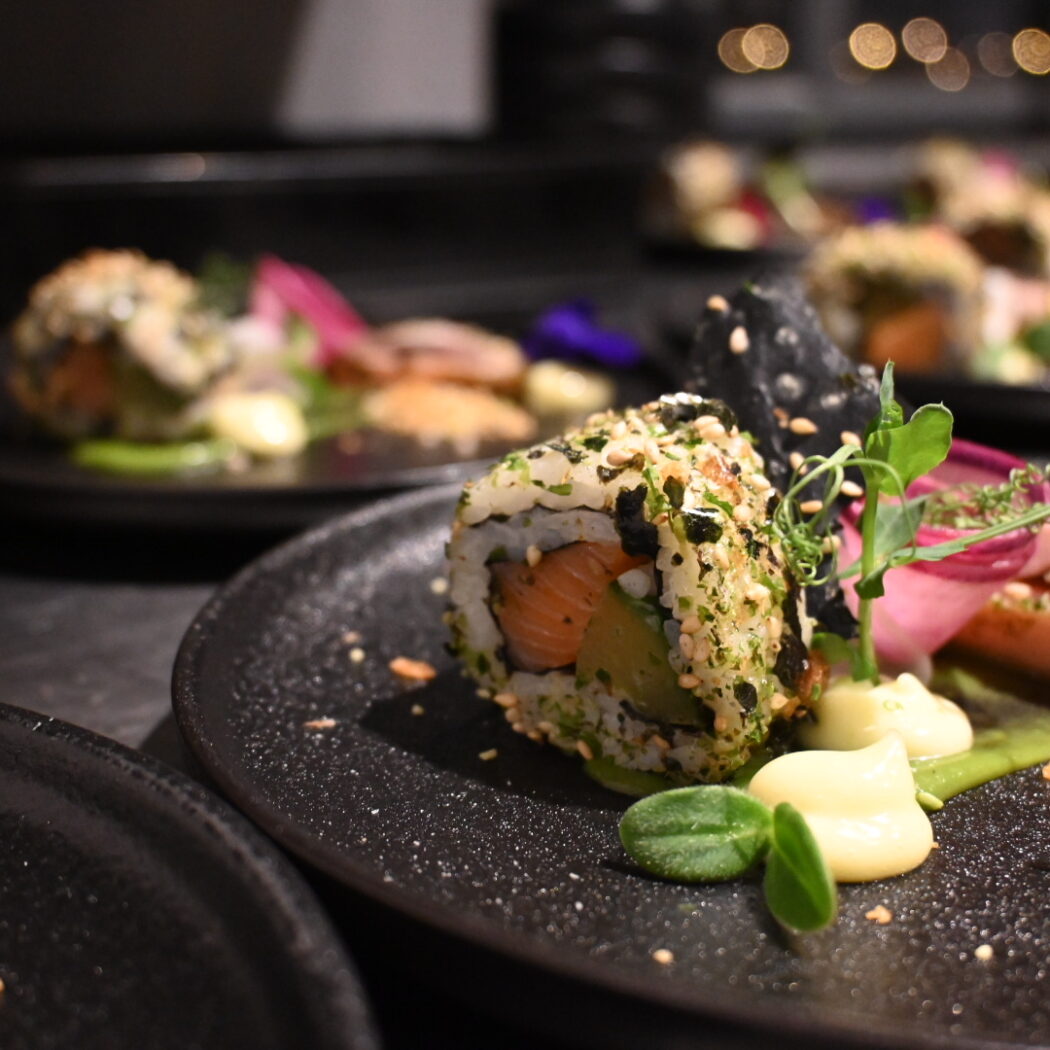 Close-up van een sushirol met zalm en avocado, bestrooid met kruiden, naast seared tonijn op een donker bord.