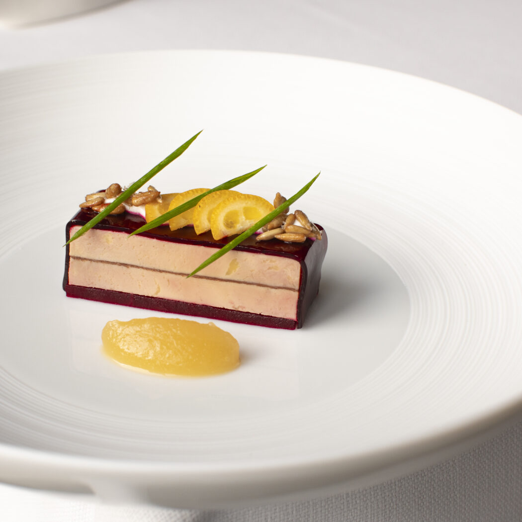 Luxe gelaagde terrine met donkerrode basis, romige vulling, citroen, pitten en bieslook op een wit bord met gele saus.
