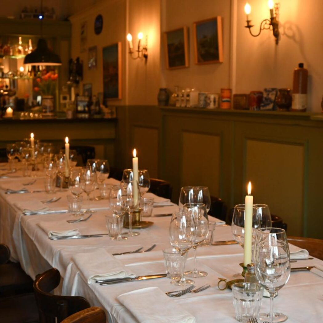 Feestelijk gedekte lange eettafel met brandende kaarsen en glazen in een sfeervol restaurant.